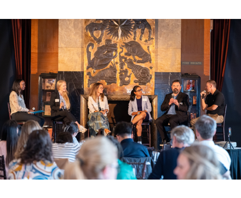 Panel of six diverse speakers on stage with an ornate brown and gold relief behind them.