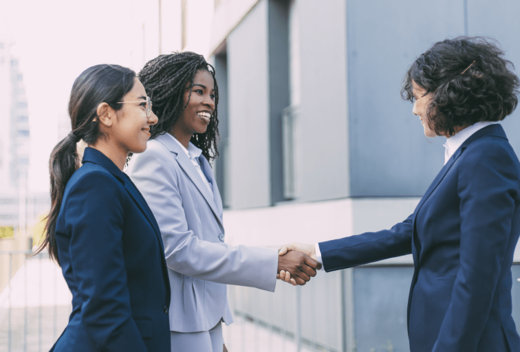 Interracial business partners greeting each other near office building. Business women wearing office suits, shaking hands with each other outside in city. Cooperation concept