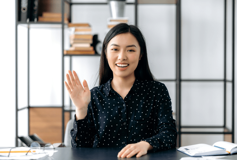 Webcam headshot of a positive pretty, smart confident young asian woman, freelancer or manager, sits at work desk, talking with colleagues or friends via video link, waving hand, smiling friendly