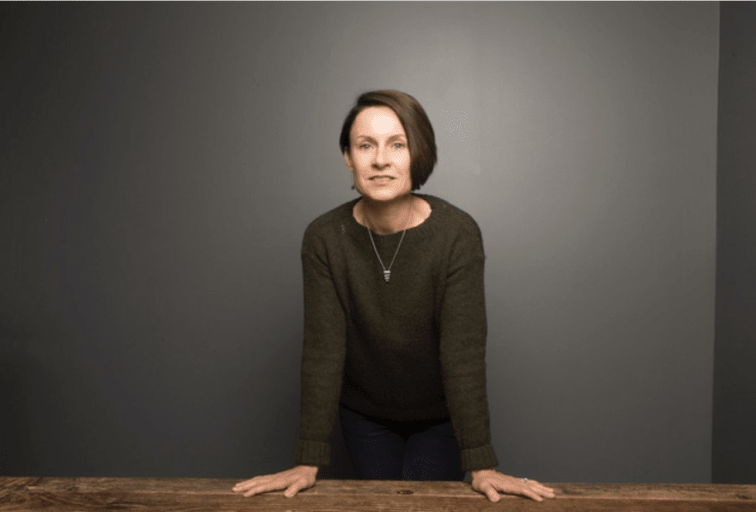 Camille Renshaw leaning on a wooden table or desk, facing the camera.