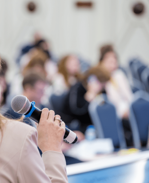 Back of anonymous elegant businesswoman sitting in chair on stage in front of audience and speaking in microphone