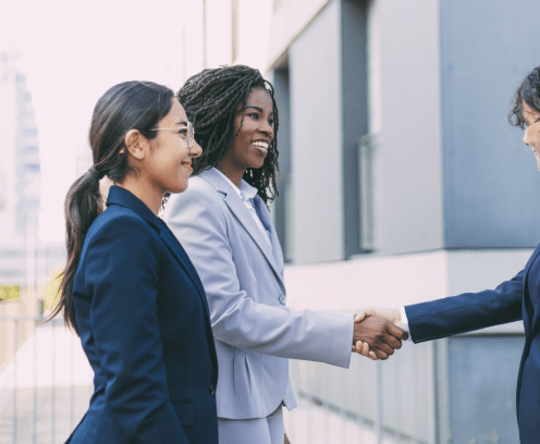 Interracial business partners greeting each other near office building. Business women wearing office suits, shaking hands with each other outside in city. Cooperation concept