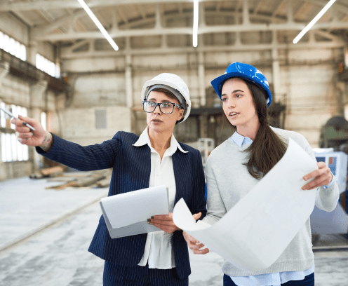 Portrait of two female engineers wearing hardhats standing in spacious workshop of modern plant pointing away and holding construction plans