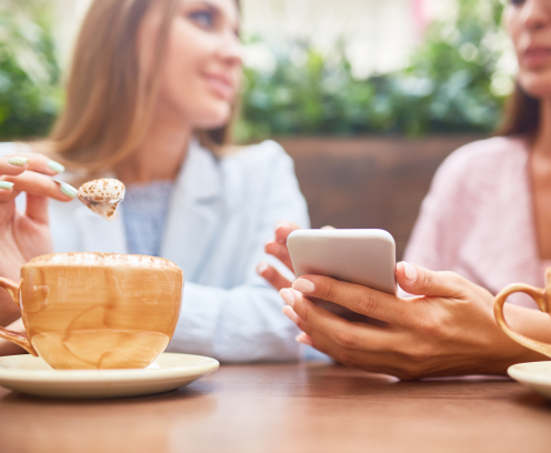 Portrait of two young women enjoying coffee at cafe table and chatting, focus on elegant female hand holding smartphone