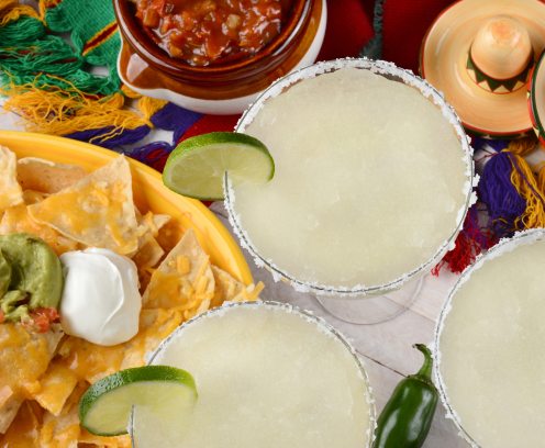 High angle view of three margarita cocktails surrounded by nachos, chips and salsa on a bright Mexican, table cloth. Horizontal format. Perfect for Cinco de Mayo projects.