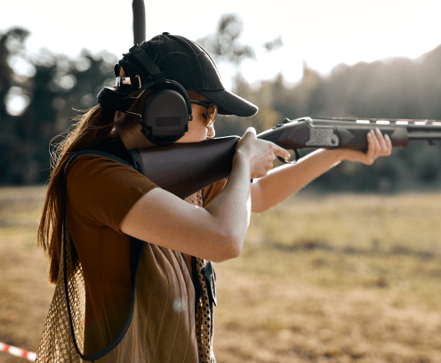 Young caucasian female on tactical gun training classes. Woman with weapon, wearing cap, protective headphones and eyeglasses. Outdoor Shooting Range. At Sunny evening at summer. Rear view
