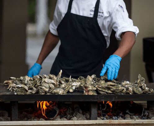 Chef with blue gloves tending to a pile of oysters roasting on a grill.