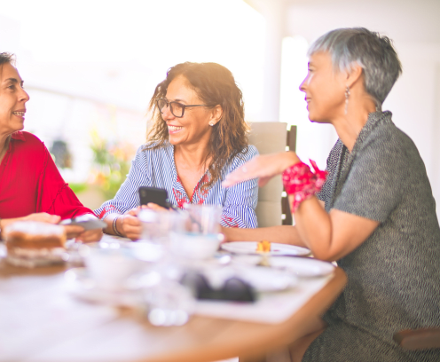 Meeting of middle age women having lunch and drinking coffee. Mature friends smiling happy using smartphone at home on a sunny day