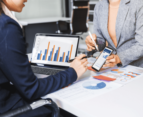 Two female business professionals in suits sit at desk, engaged in strategic discussion. They analyze financial planning, risk management, corporate growth while reviewing reports and market trends.