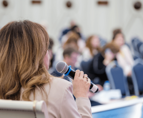 Back of anonymous elegant businesswoman sitting in chair on stage in front of audience and speaking in microphone