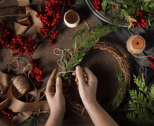 Table with hands decorating a wreath with pine, berries and gold ribbons.