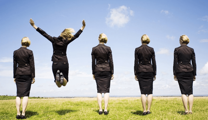 five businesswomen on grass with blue sky with one jumping in air