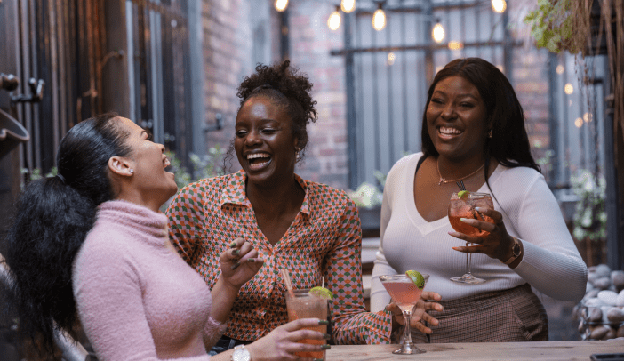 Medium shot of three female friends drinking cocktails and dancing together on an outdoor terrace in the North East of England. They are laughing a having fun together.