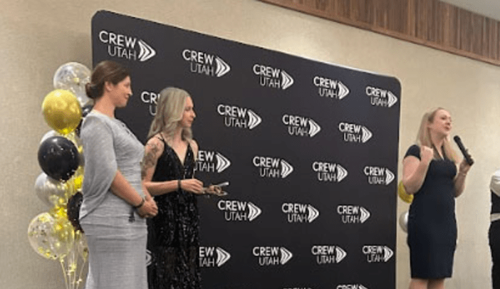 Female speaker in front of CREW Utah Banner with two female award recipients