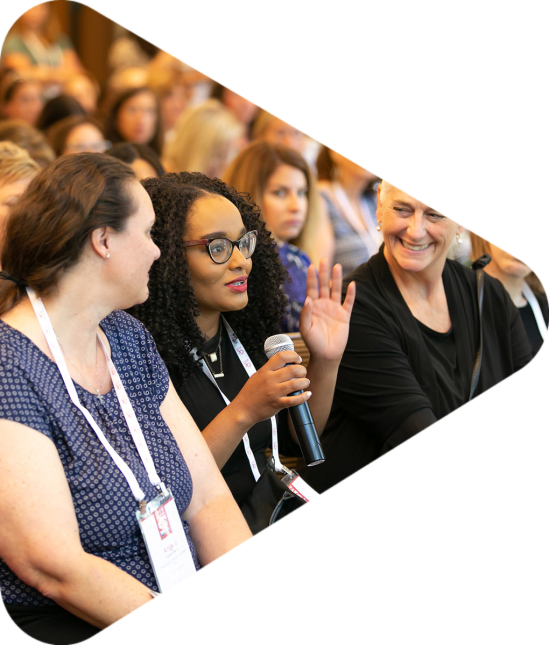 woman holding microphone and talking whilst sitting in row of a convention crowd