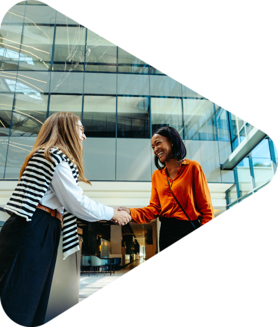 A welcoming scene of two professional women exchanging a friendly handshake in a modern office lobby. The spacious glass surroundings evoke a sense of openness and collaboration.