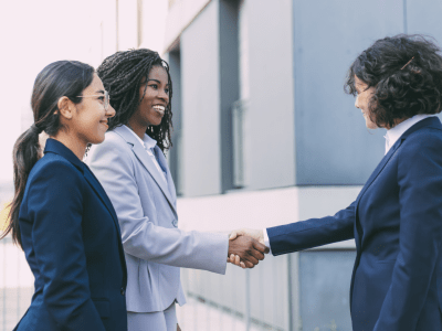 Interracial business partners greeting each other near office building. Business women wearing office suits, shaking hands with each other outside in city. Cooperation concept