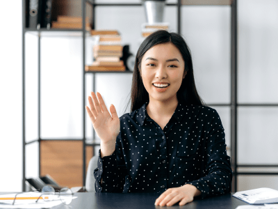 Webcam headshot of a positive pretty, smart confident young asian woman, freelancer or manager, sits at work desk, talking with colleagues or friends via video link, waving hand, smiling friendly