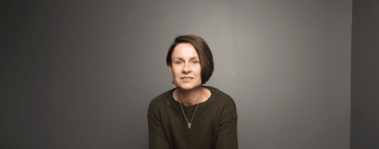 Camille Renshaw leaning on a wooden table or desk, facing the camera.