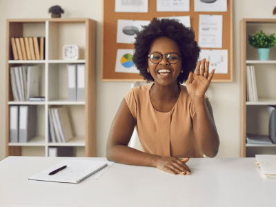Video or online lesson. African american woman waving hello smiling looking at camera sitting at office desk. Excited businesswoman teacher lecturer recording educational webinar, greeting students
