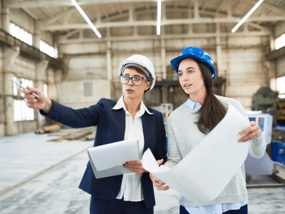 Portrait of two female engineers wearing hardhats standing in spacious workshop of modern plant pointing away and holding construction plans