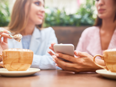 Portrait of two young women enjoying coffee at cafe table and chatting, focus on elegant female hand holding smartphone