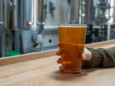 Women holding pint of beer on counter at front of fermentation tanks and some brewery equipment