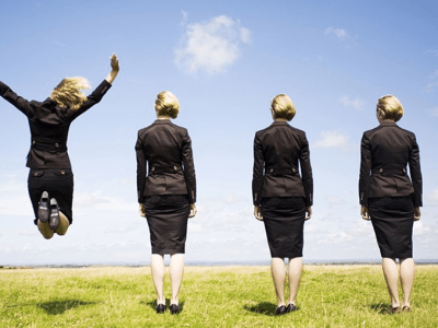 five businesswomen on grass with blue sky with one jumping in air