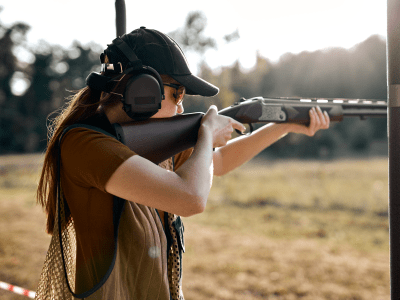 Young caucasian female on tactical gun training classes. Woman with weapon, wearing cap, protective headphones and eyeglasses. Outdoor Shooting Range. At Sunny evening at summer. Rear view