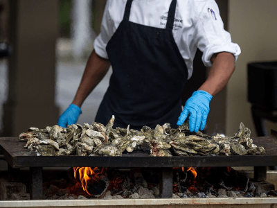 Chef with blue gloves tending to a pile of oysters roasting on a grill.