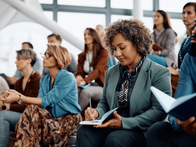 Mature entrepreneur writing in her notebook while attending a business seminar at conference hall.