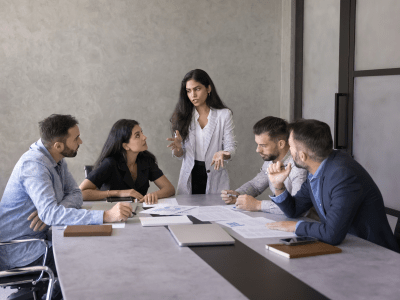 Young Indian business project manager woman presenting marketing reports, sales data to team, speaking to colleagues, standing at large meeting table. Diverse group analyzing financial research