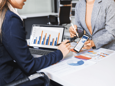 Two female business professionals in suits sit at desk, engaged in strategic discussion. They analyze financial planning, risk management, corporate growth while reviewing reports and market trends.