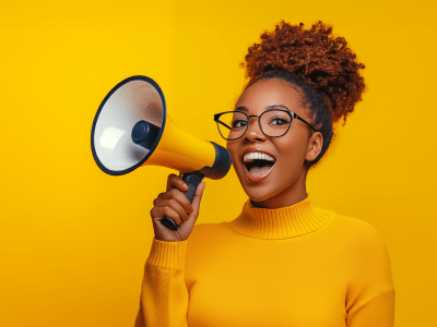 Happy young Black woman in yellow sweater,  announcing sale offers through a megaphone, isolated on a yellow background with copy space. Screaming into the megaphone. Studio portrait.