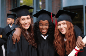 Happy group of mature students on graduation day embracing with each other. Three best girl friends in academic gowns and caps hugging in front of the camera