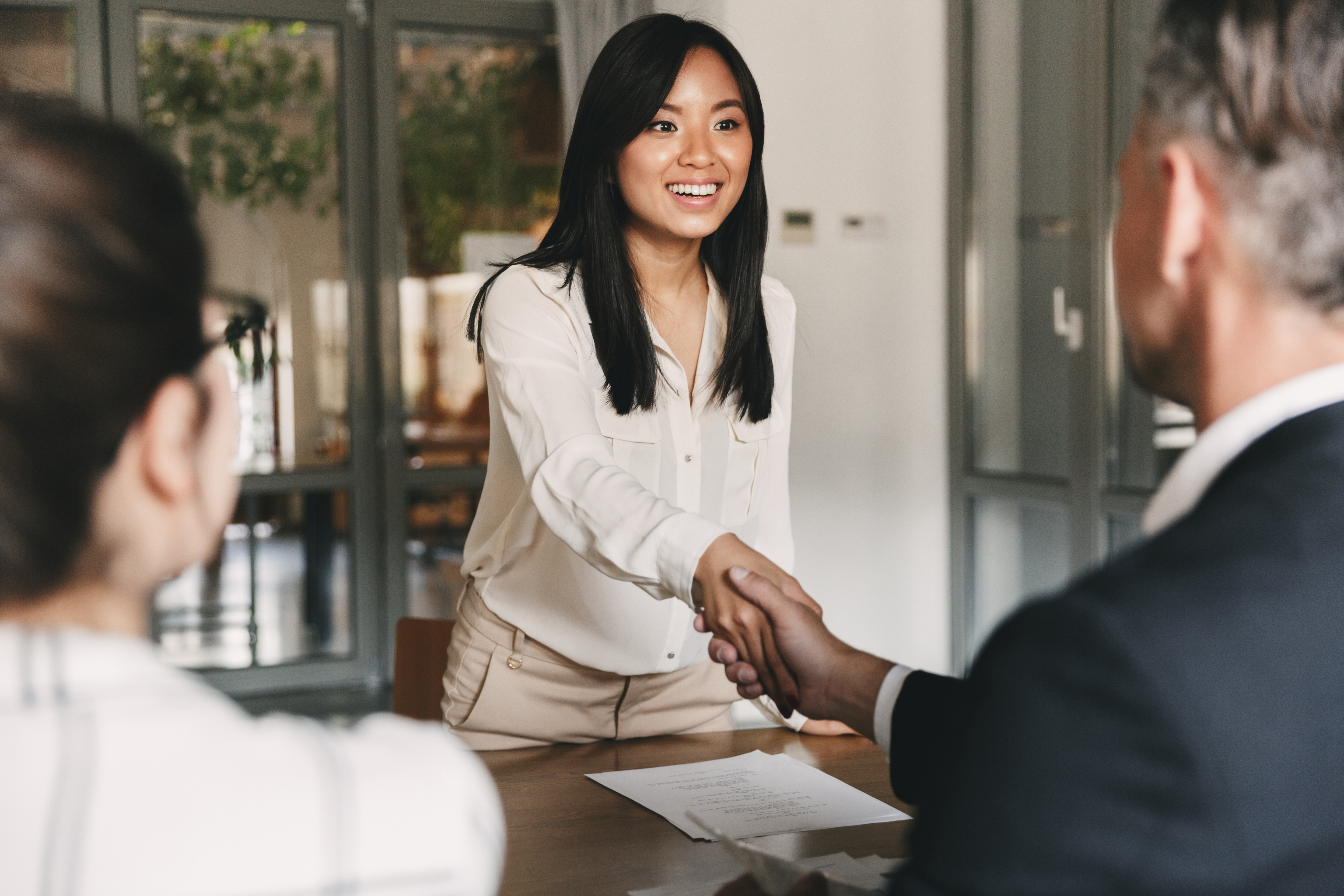 Business, career and placement concept - image from back of two employers sitting in office and shaking hand of young asian woman after successful negotiations or interview